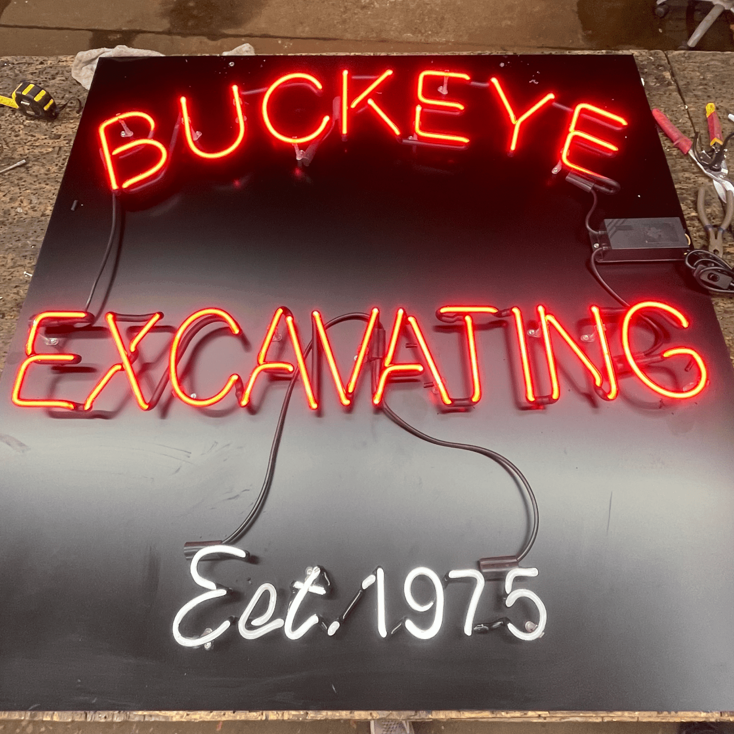 Red and white neon tubing spelling BUCKEYE EXCAVATING Est. 1975 lit on a shop workbench