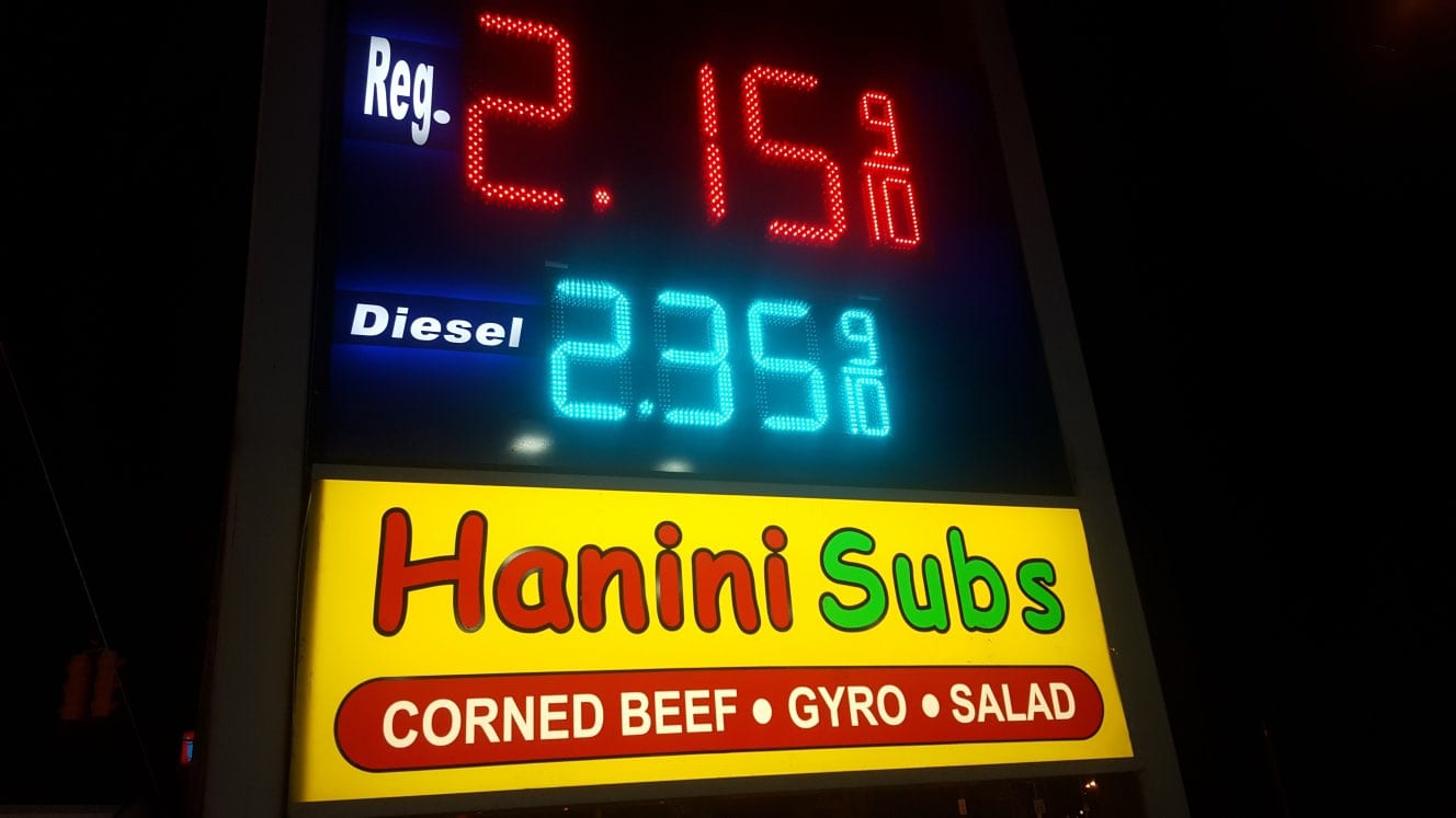 Lit pylon sign at night with LED fuel prices above a yellow Hanini Subs panel