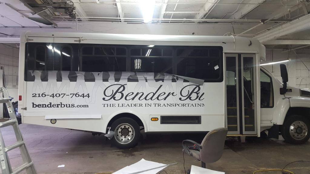 Black Bender Bus vinyl lettering being applied to the side of a white shuttle bus indoors