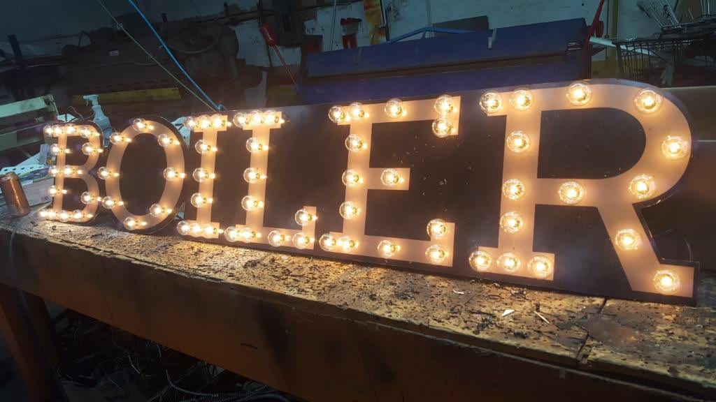 Sheet metal marquee letters spelling BOILER with lit bulbs on a shop workbench