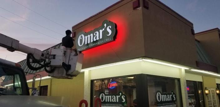Worker in a bucket lift beside a red halo-lit Omar's sign at dusk