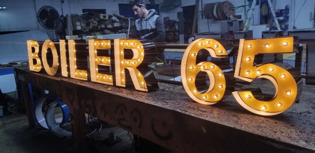 Bulb-lit marquee letters spelling BOILER 65 on a shop bench with a fabricator working behind