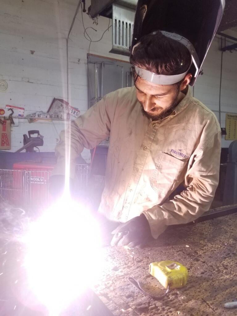 Shop worker in a welding helmet at a steel bench as a bright arc flares