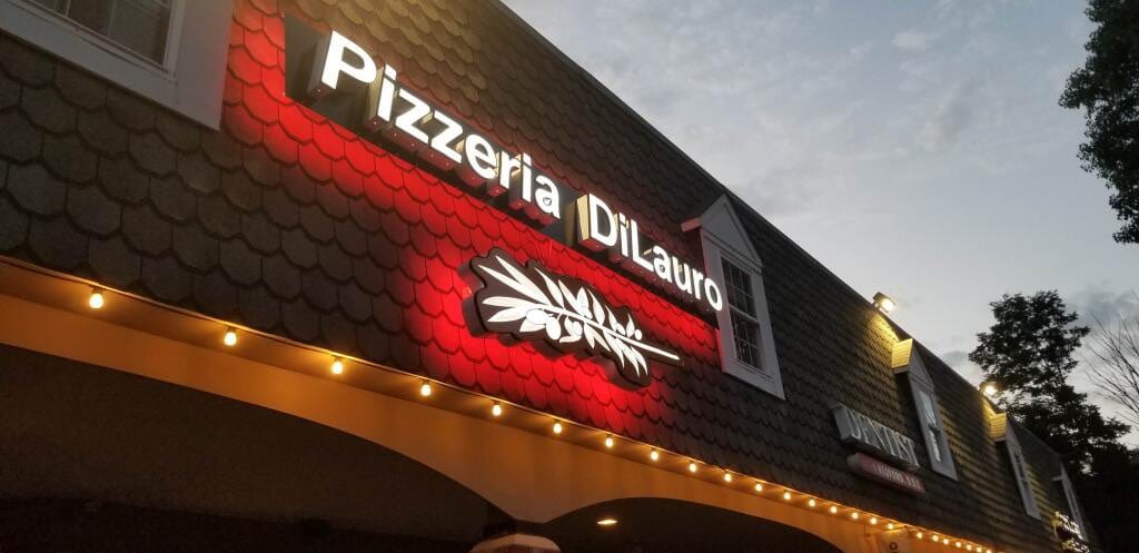 Lit Pizzeria DiLauro letters and wheat logo glowing red on a shingled facade at dusk