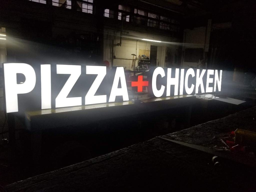 Lit PIZZA + CHICKEN channel letters resting on a workbench inside the sign shop