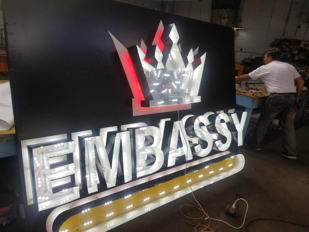 Worker in the shop beside an Embassy sign with exposed LED modules inside the letters and crown