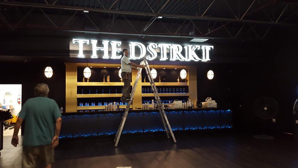 Man on a ladder working beneath lit THE DSTRKT letters above a bar back