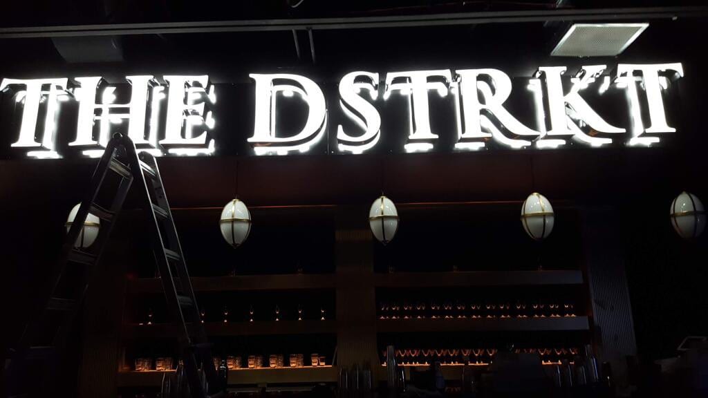 Lit THE DSTRKT letters glowing above a bar interior with a ladder alongside