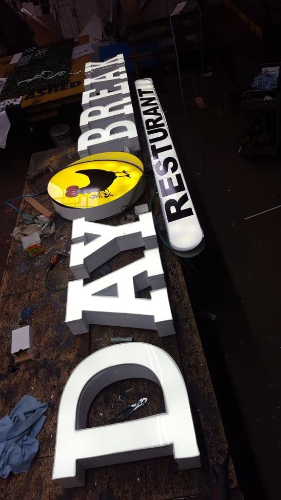 Lit DAY BREAK RESTURANT letters and an oval logo cabinet laid out on a shop bench