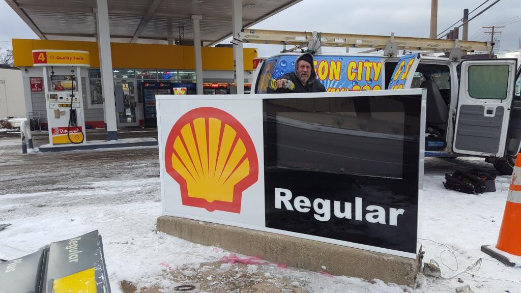 Technician working behind a Shell gas station price sign cabinet with a sign service van alongside