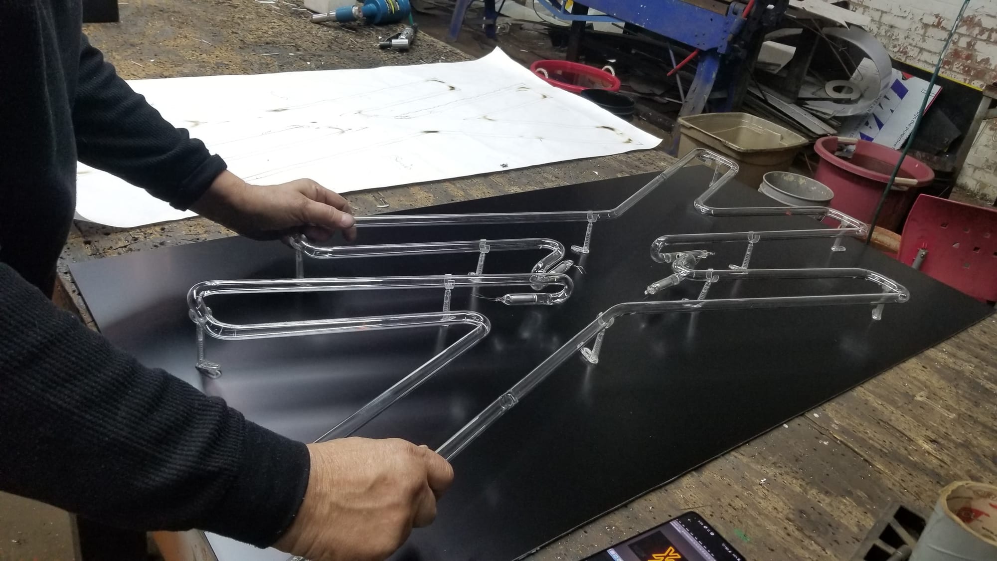 Hands laying out bent clear neon tubing letters on a black panel at a shop bench