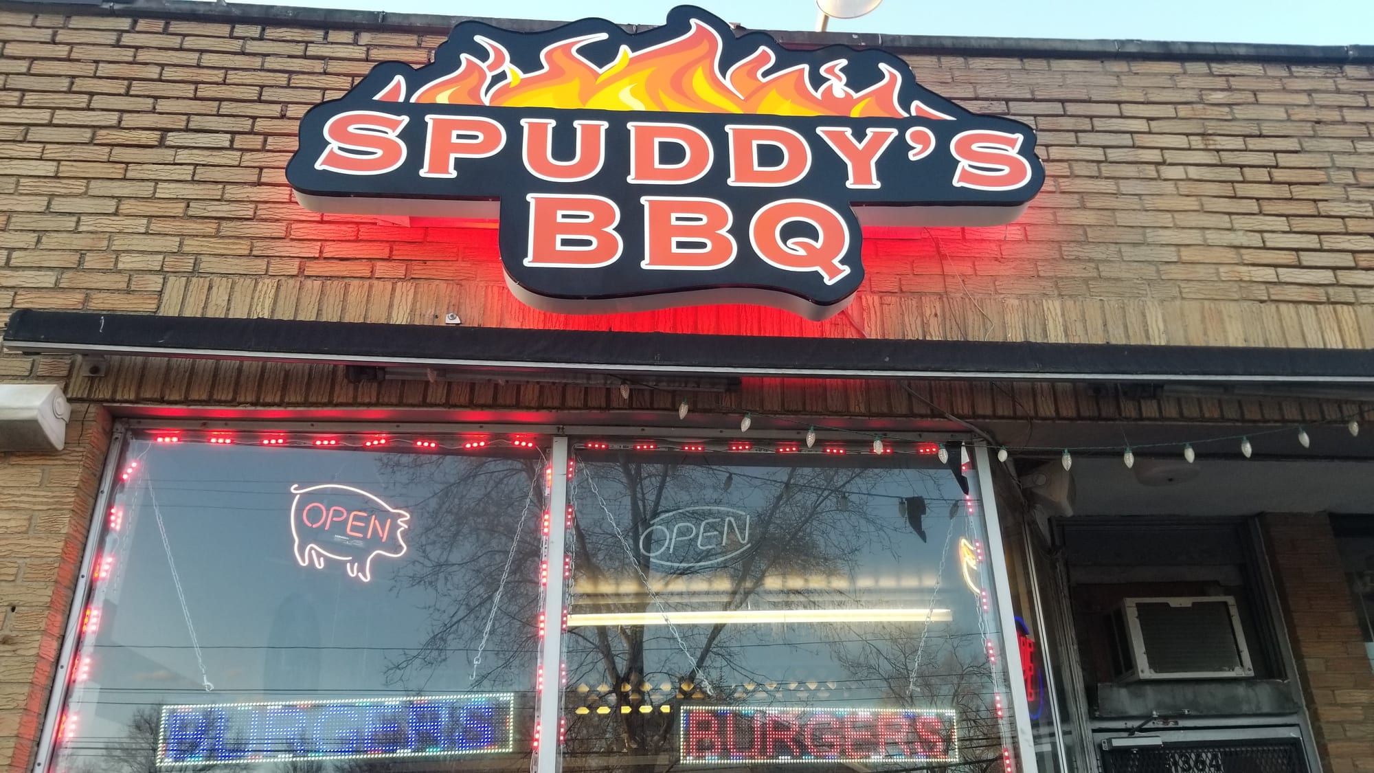Lit Spuddy's BBQ shaped cabinet sign with red halo glow on a brick storefront