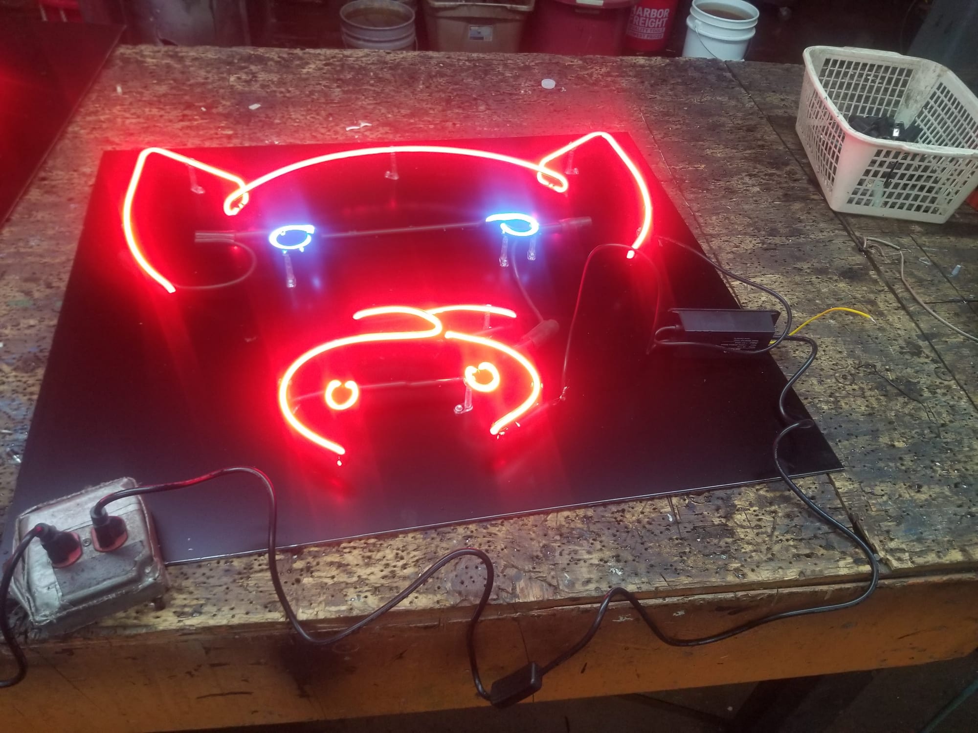 Neon pig face sign glowing red and blue on a black panel on a shop workbench