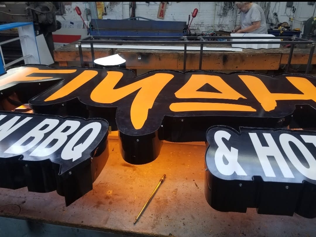 Black channel letter sections for a BBQ sign laid out on a shop bench near a worker