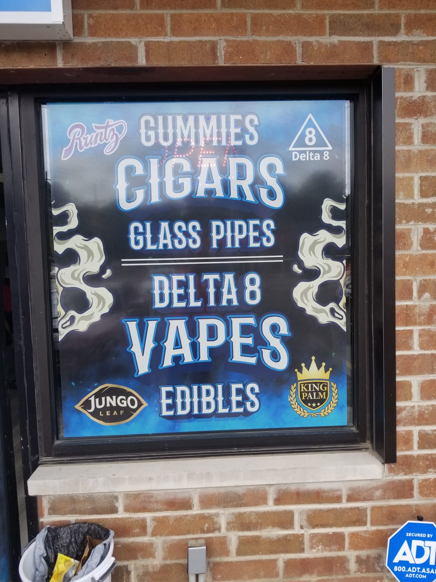 Window graphic listing cigars, glass pipes, Delta 8, vapes and edibles on a brick wall