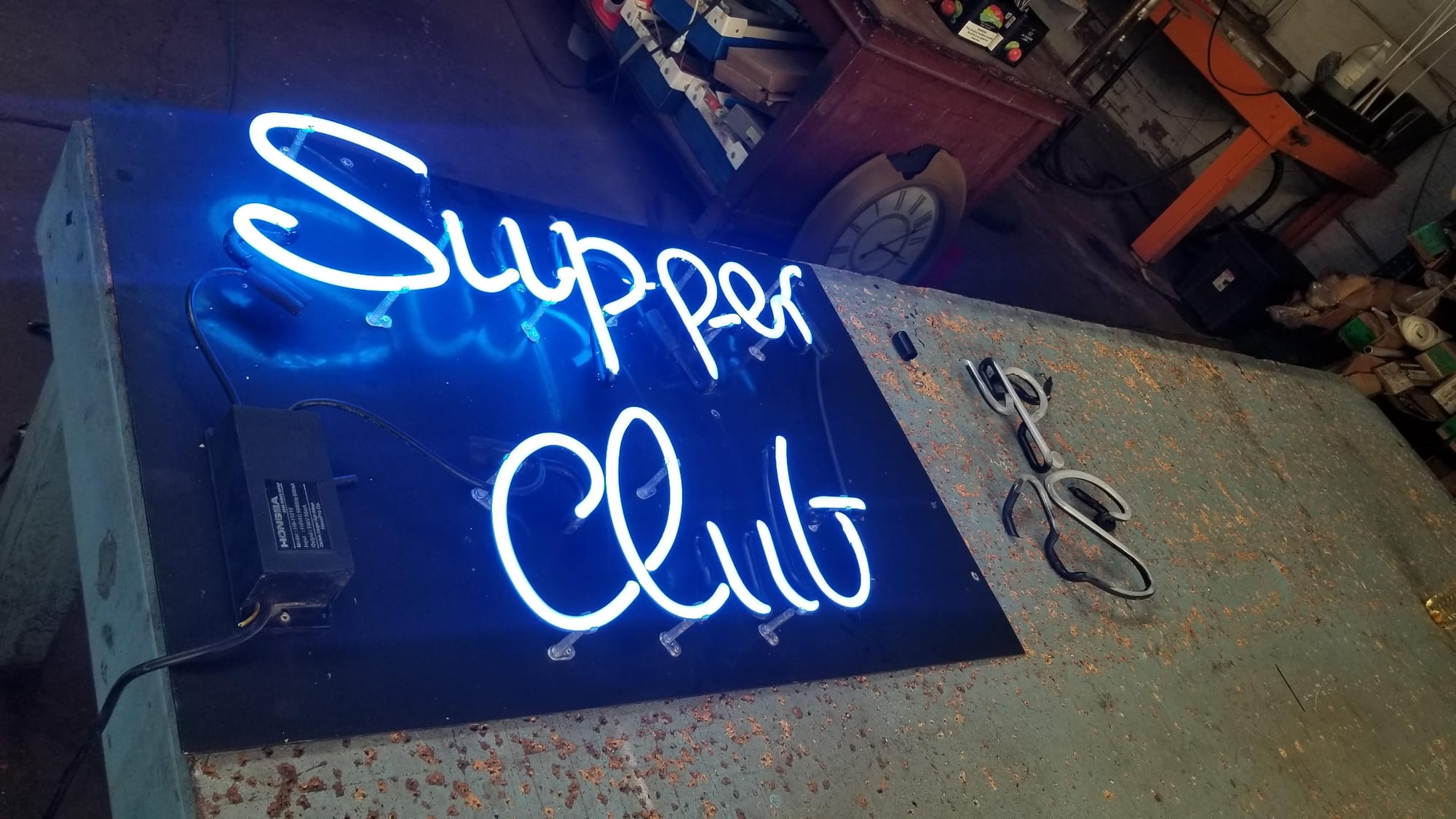 Blue neon Supper Club sign lit on a black backer panel on a shop table