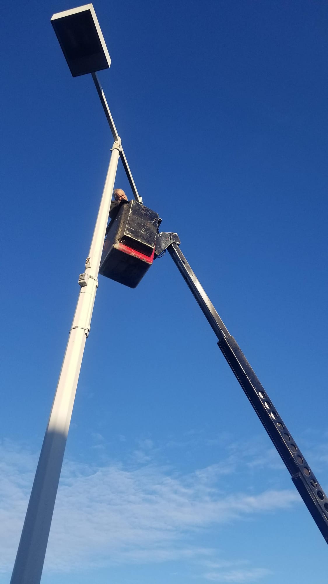 Worker in a bucket lift at the top of a parking lot light pole against blue sky