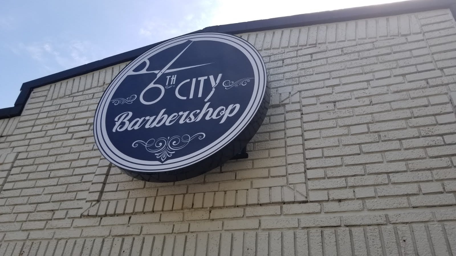 Round 6th City Barbershop cabinet sign with scissors logo on a white brick wall