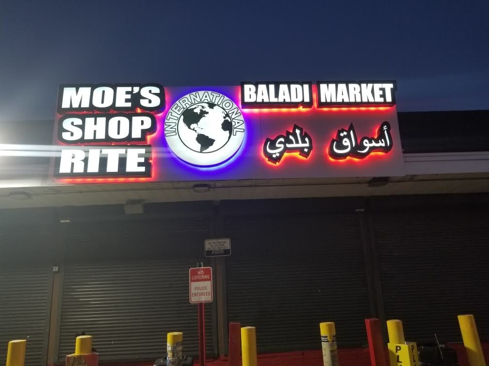 Lit MOE'S SHOP RITE and BALADI MARKET letters with a globe logo above a storefront