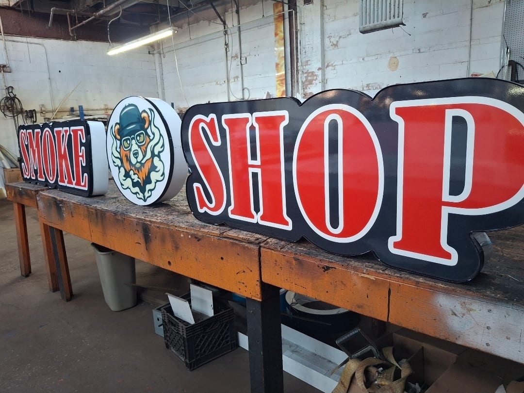 SMOKE SHOP sign faces and a bear logo cabinet resting on a workbench in the fabrication shop