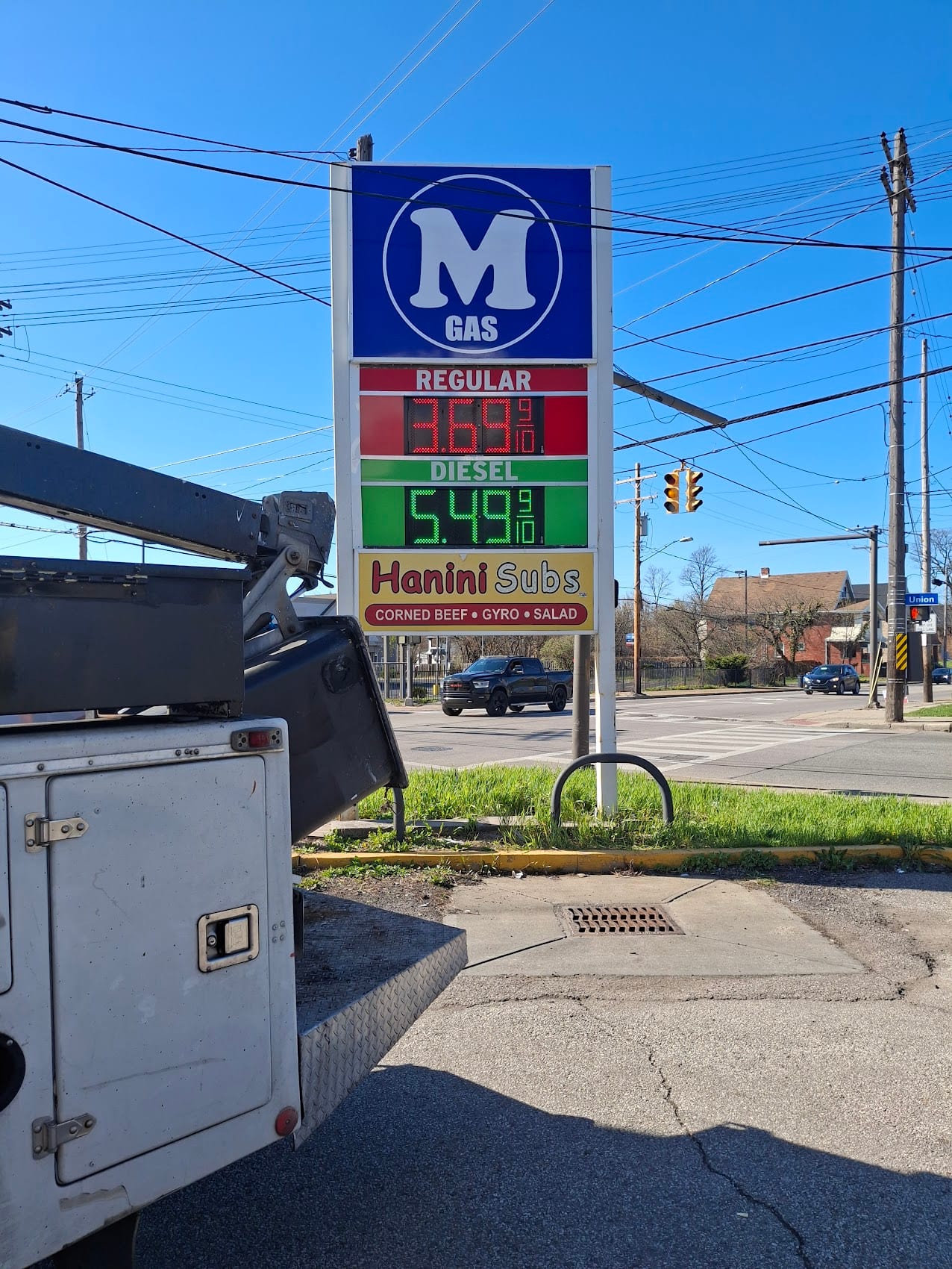 M Gas pylon sign with LED fuel prices and a Hanini Subs panel at a street corner