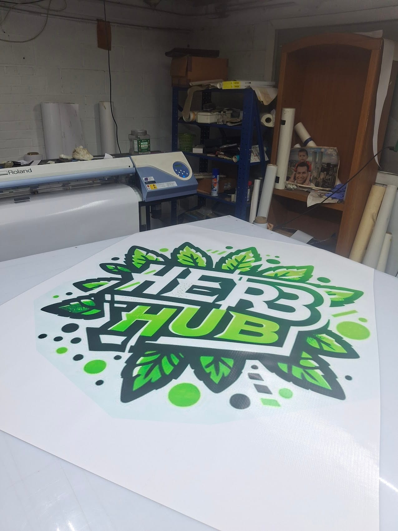 Printed Herb Hub banner laid out in the shop beside a Roland printer