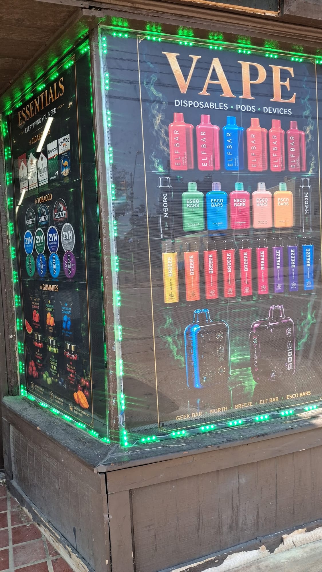 Printed VAPE and ESSENTIALS window panels on a corner storefront with green LED trim